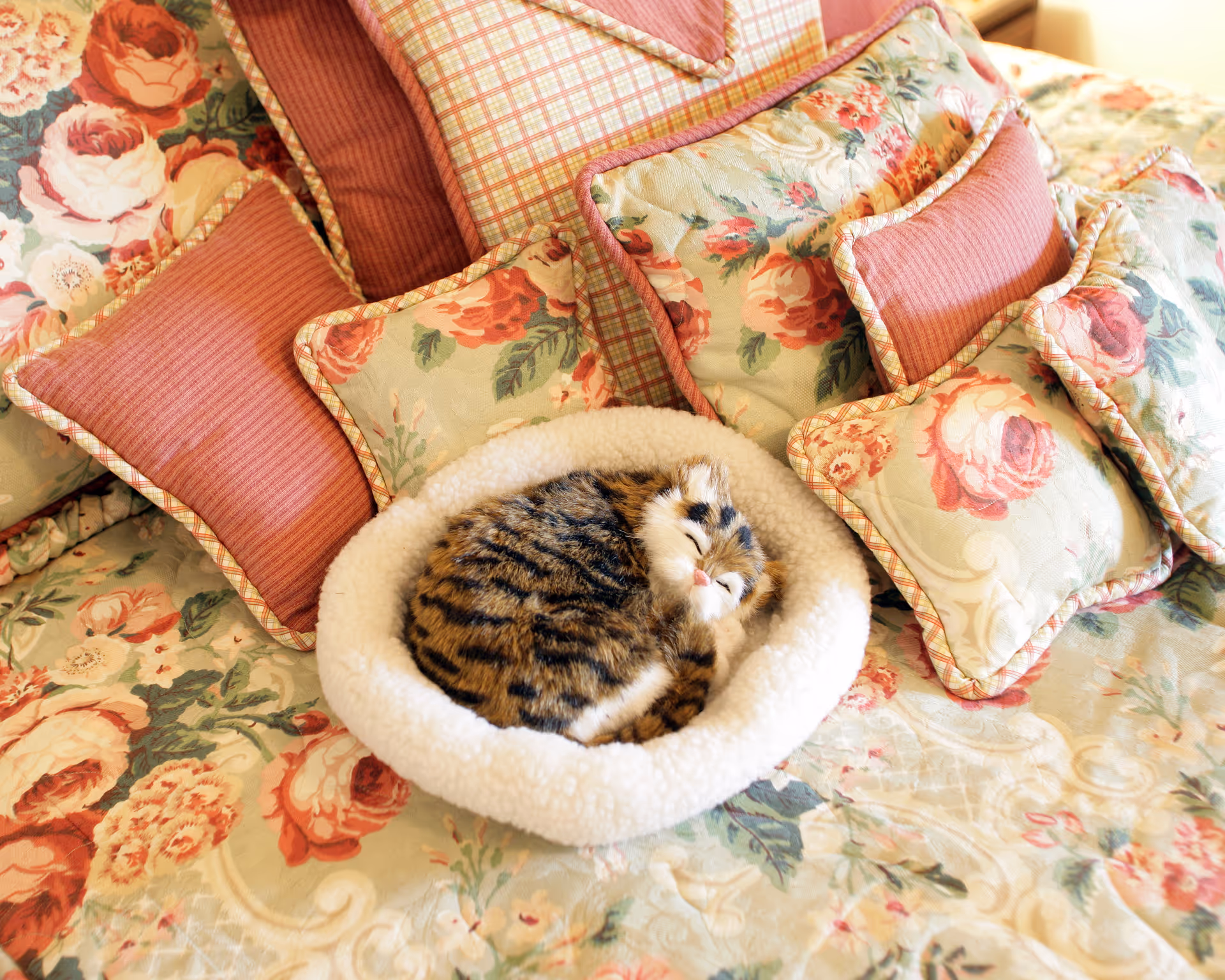 A small tabby cat curled up and sleeping in a white fluffy pet bed placed on a floral-patterned bedspread with multiple decorative pillows in shades of pink and floral designs.