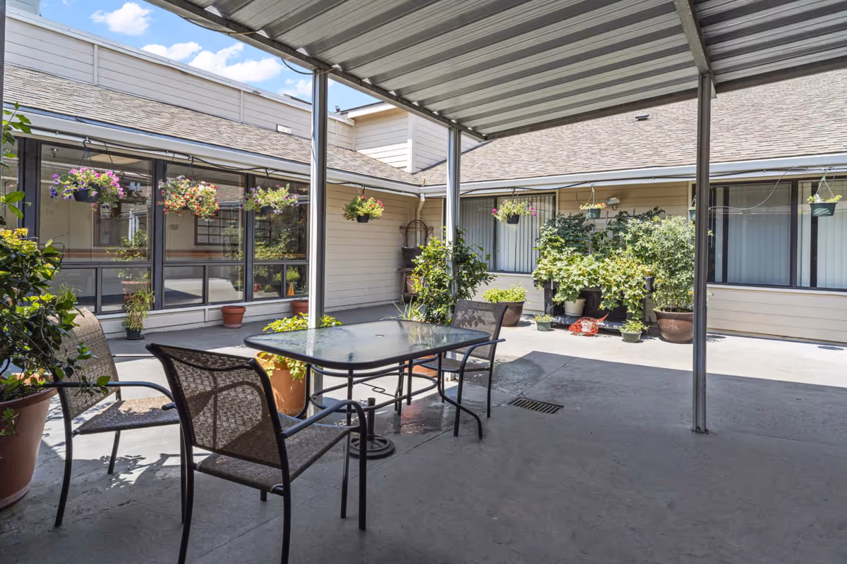 Outdoor covered patio area at Linda Vista Nursing and Rehab Center with a glass-top table and four mesh chairs. The patio is surrounded by potted plants and hanging flower baskets, with beige building walls and windows in the background under a partly cloudy sky.