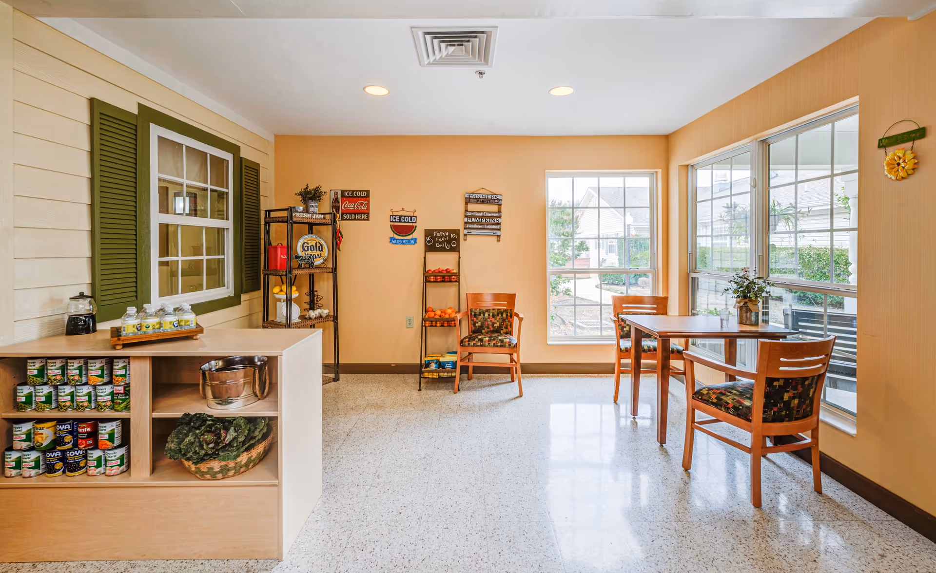 A bright and clean dining area in a senior living facility with a small wooden table and three chairs with patterned cushions near large windows. There is a counter with canned goods and a basket of vegetables, a metal rack with fruit, and vintage-style signs on the wall. The walls are painted a warm beige color.