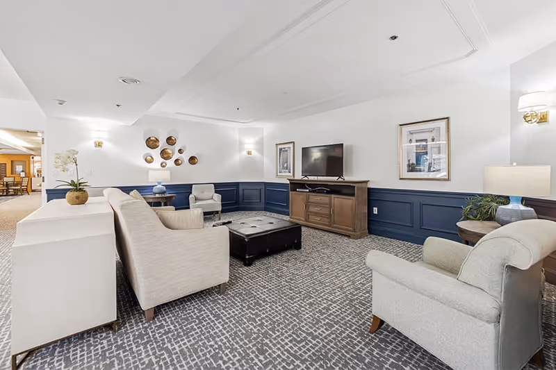 A cozy living room area in a senior living facility with beige upholstered armchairs and a sofa arranged around a dark tufted ottoman. The walls are white with navy blue wainscoting, decorated with framed artwork and a wall-mounted TV on a wooden console. The carpet has a geometric pattern, and there are table lamps and wall sconces providing warm lighting.