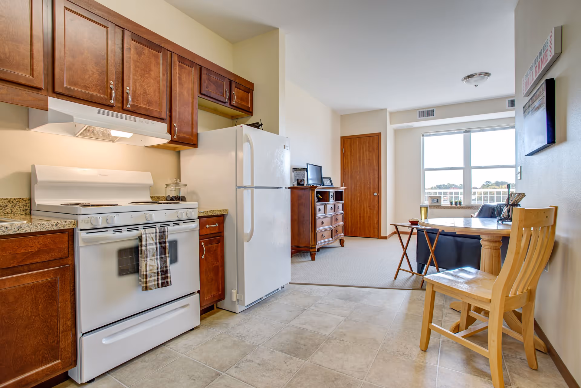 Interior view of a senior living facility kitchen and dining area. The kitchen features wooden cabinets, a white stove with a towel hanging on the handle, and a white refrigerator. Adjacent to the kitchen is a small dining area with a wooden chair and a round table. In the background, there is a living area with a wooden dresser, a TV, and a large window letting in natural light.