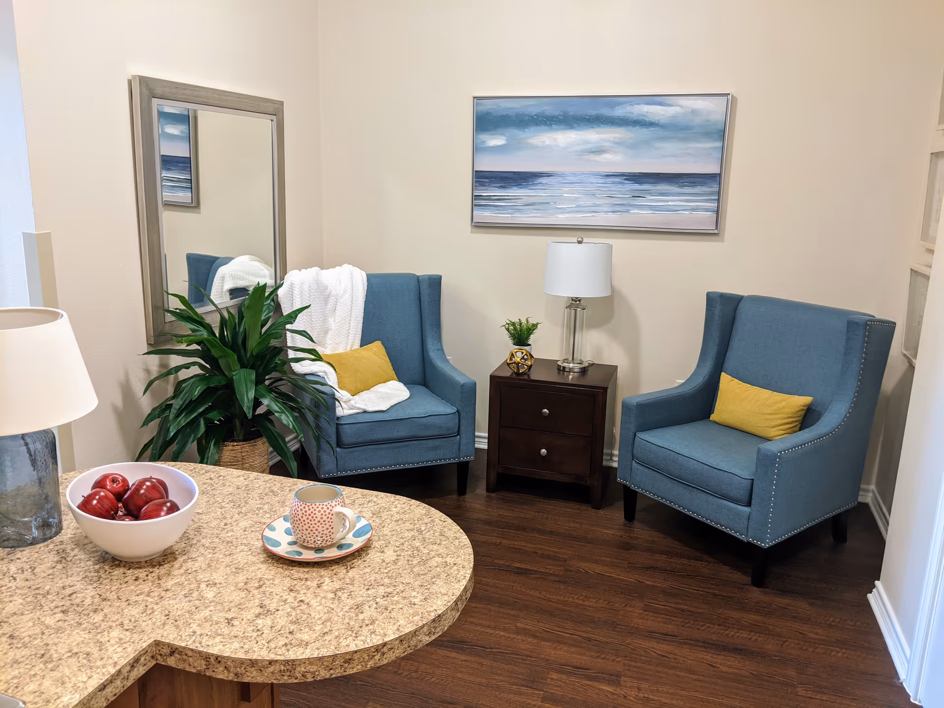 Small sitting area with two blue armchairs, a side table and lamp, a plant, wall art, and a countertop with a bowl of apples and a cup.
