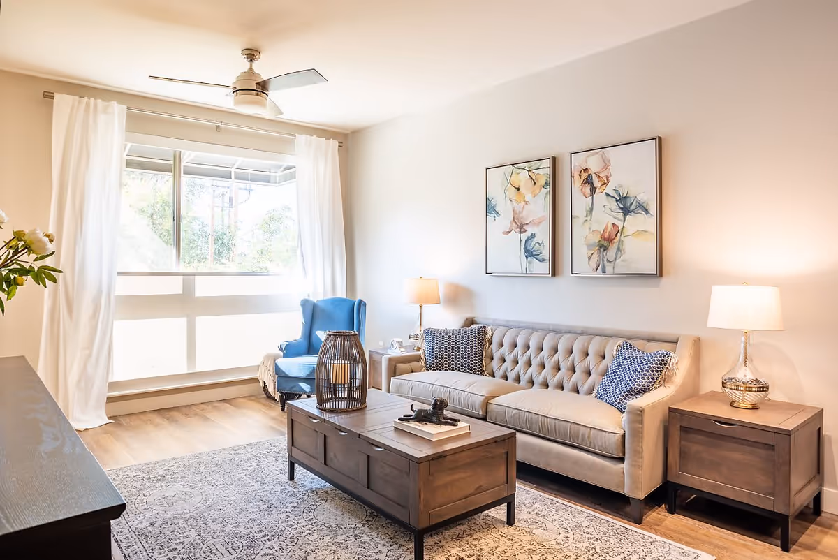A bright and cozy living room with a large window covered by white curtains, a beige tufted sofa with two patterned pillows, a blue armchair, two framed floral paintings on the wall, two side tables with lamps, a wooden coffee table with decorative items, and a ceiling fan.