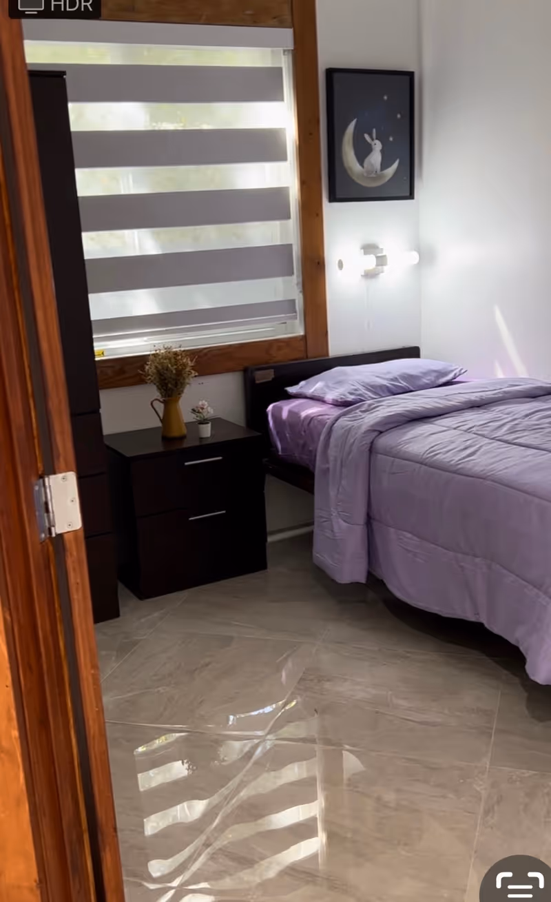 A small bedroom with a single bed covered in light purple bedding. Next to the bed is a dark wooden nightstand with two drawers, holding a small vase with dried flowers and a small potted plant. A window with horizontal blinds is above the nightstand, and a framed picture of a rabbit sitting on a crescent moon hangs on the white wall above the bed. The floor is tiled with a glossy finish.