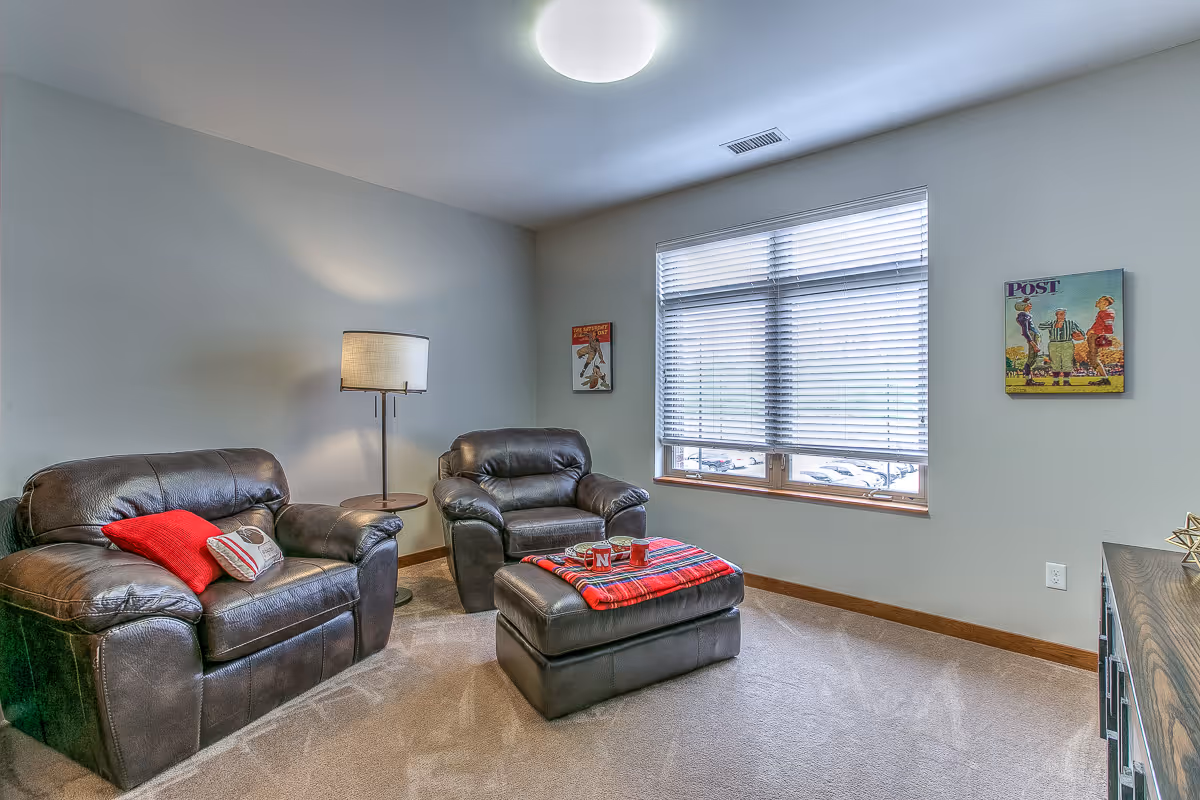 A cozy living room with two dark leather armchairs, a matching ottoman with a red plaid blanket and mugs on top, a floor lamp, and a window with blinds. The walls are light gray with two framed vintage-style posters.