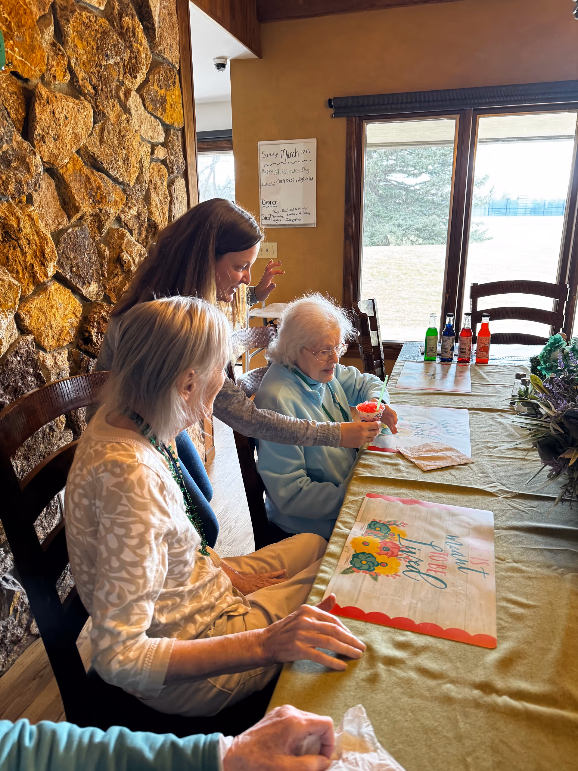 Three women sitting at a table in a room with a stone wall and large window. Two elderly women are seated, one wearing a light blue sweater and the other a white patterned top, while a younger woman stands behind them, smiling and assisting with a red drink. The table is covered with a green tablecloth and has placemats with floral designs and text. Bottles of colorful syrup or drink mixers are on the table near the window.