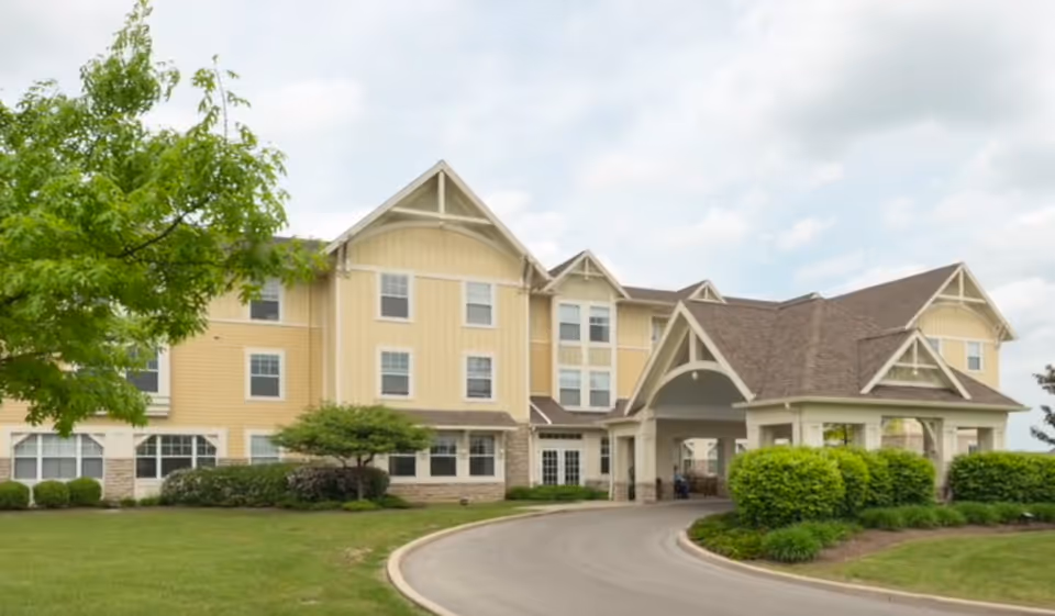 Yellow multi-story senior living building with a covered porte-cochère entrance, curved driveway and landscaped lawn.