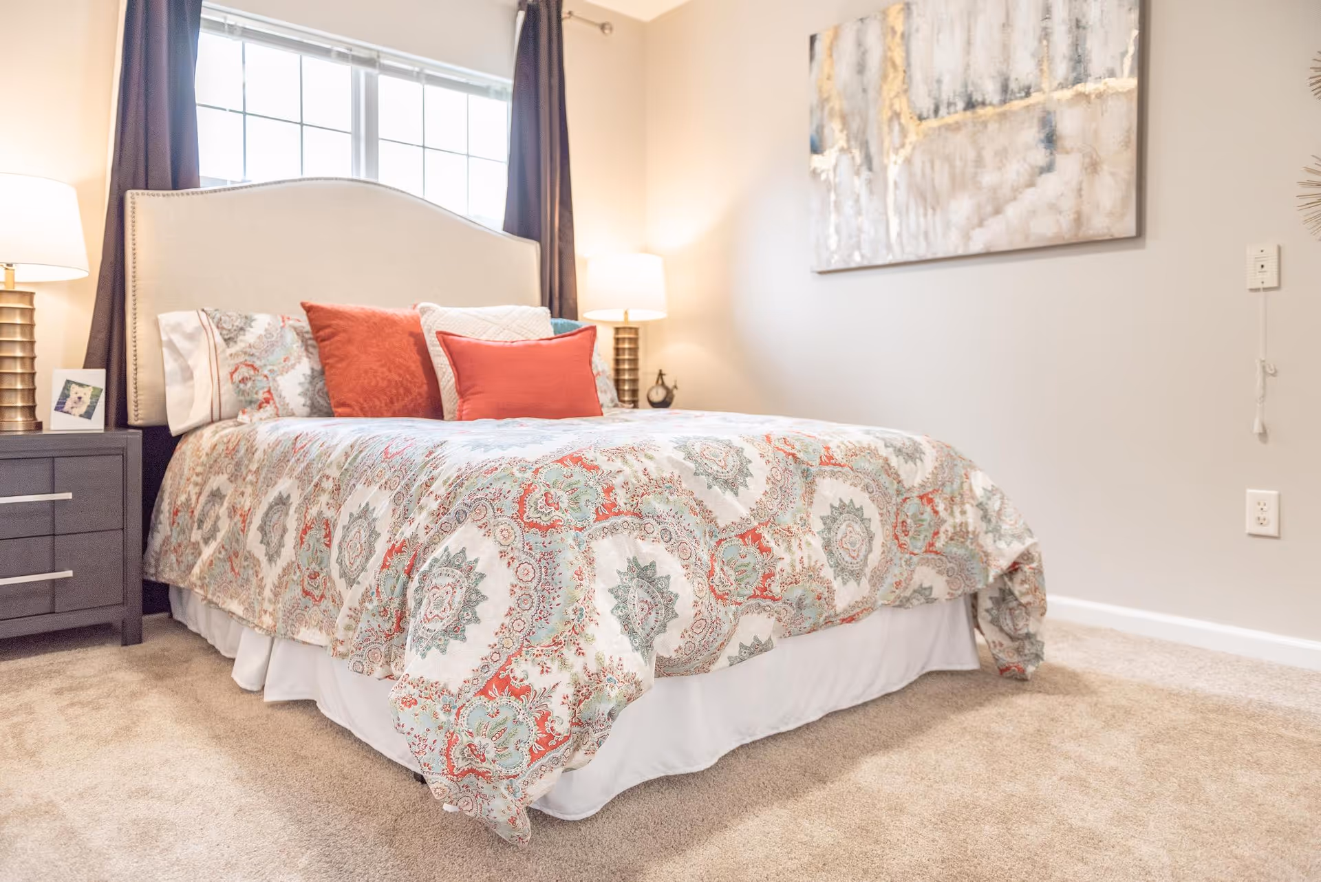 A neatly made bed with a patterned bedspread and multiple pillows in shades of red and white, positioned against a wall with a large window behind it. There are two bedside tables with lamps on either side of the bed, and a piece of abstract artwork hanging on the adjacent wall. The room has beige carpeting and light-colored walls.