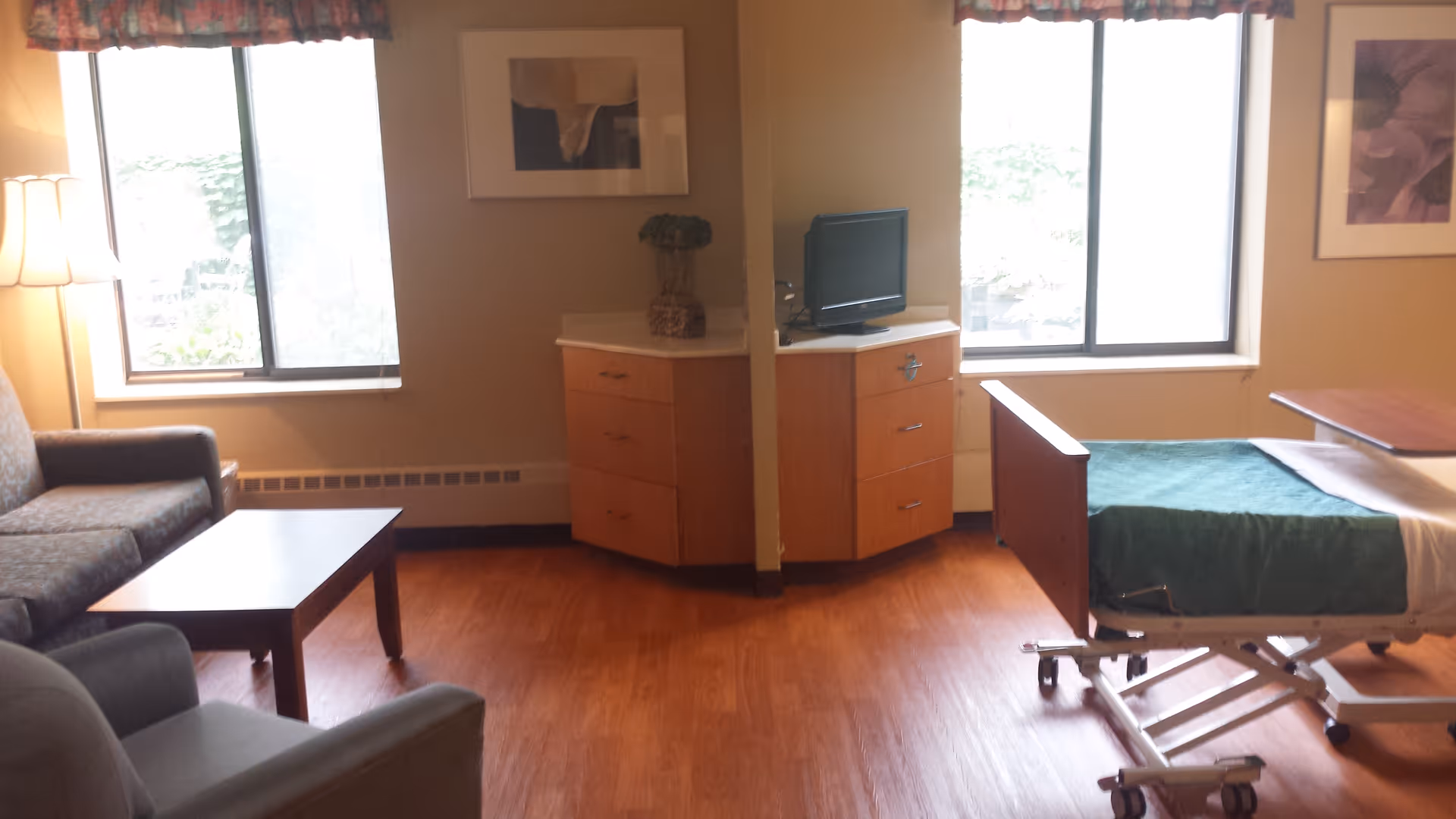 A room with a hospital-style bed with green bedding, a small TV on a corner cabinet, two windows with floral valances, a couch, an armchair, a coffee table, and framed artwork on the walls.