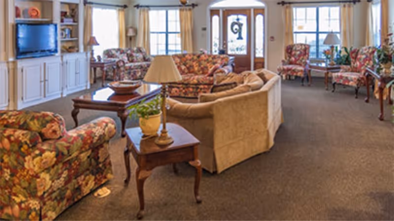 Well-lit communal living room with sofas and floral armchairs arranged around coffee tables and a television.