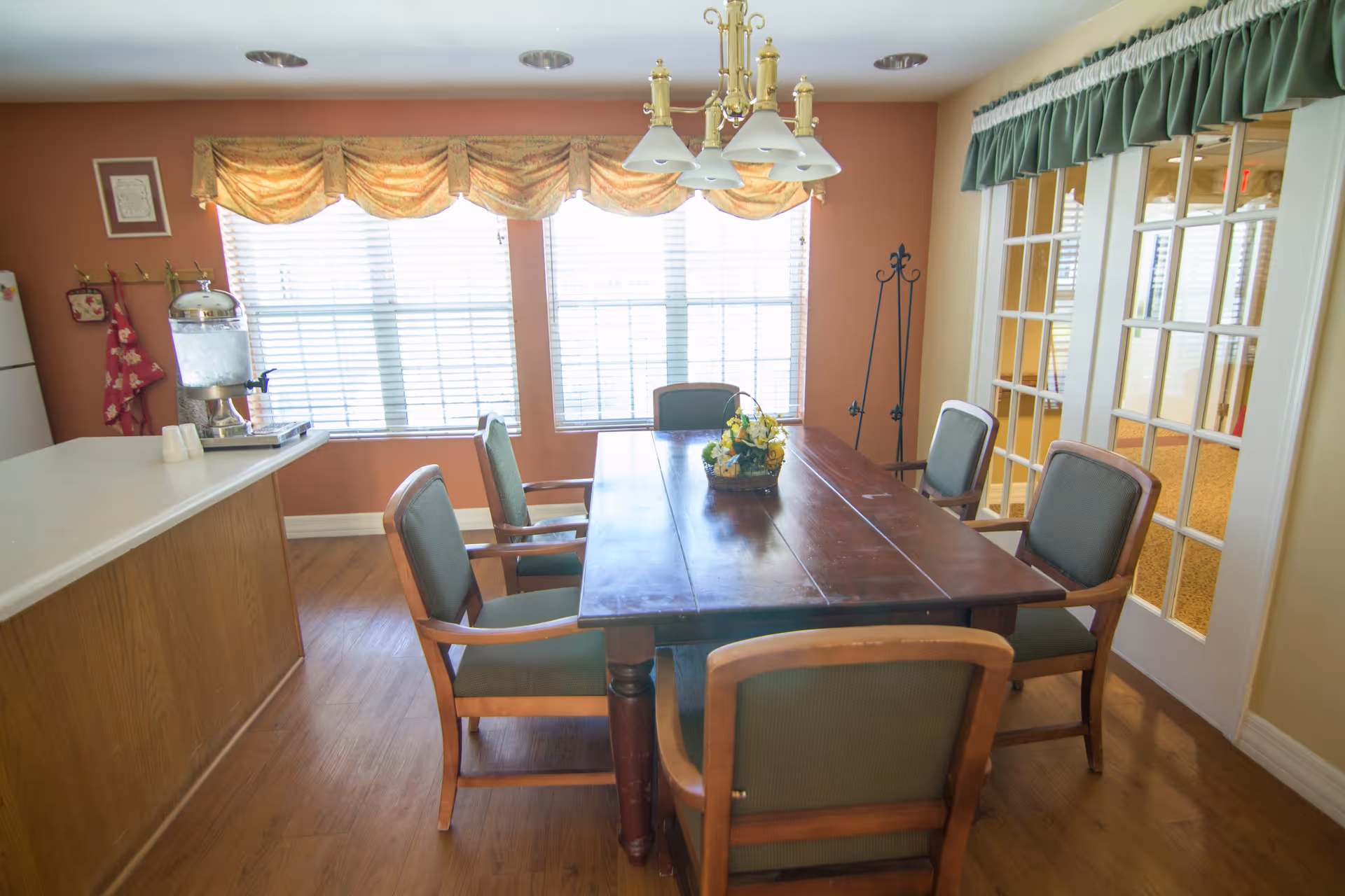 A dining room with a wooden table surrounded by six chairs with green cushions. There is a floral centerpiece on the table. The room has large windows with blinds and decorative valances, a chandelier hanging above the table, and a counter with a water dispenser on the left side. French doors with glass panes are on the right side.