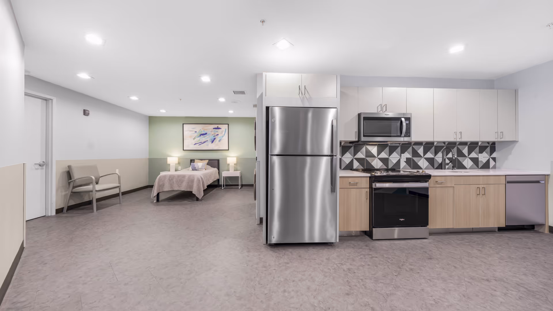 A spacious, modern room featuring a kitchenette with stainless steel appliances including a refrigerator, microwave, stove, and dishwasher. The kitchenette has light wood lower cabinets and white upper cabinets with a geometric black, white, and gray tile backsplash. In the background, there is a bed with a light pink bedspread, two bedside tables with lamps, a framed abstract painting on a green accent wall, and a beige chair near a closed door.