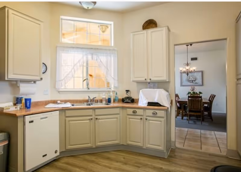A kitchen area with cream-colored cabinets and a wooden countertop. There is a window with a sheer curtain above the sink, and various items such as soap dispensers and a covered appliance on the counter. Through an open doorway, a dining room with a wooden dining table, chairs, a chandelier, and a framed picture on the wall is visible.
