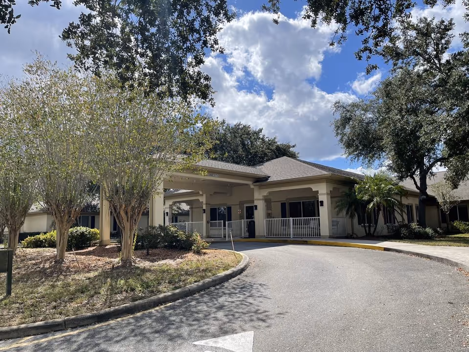 Front entrance of a single-story senior living facility with a covered drop-off driveway, landscaping, and trees under a partly cloudy sky.