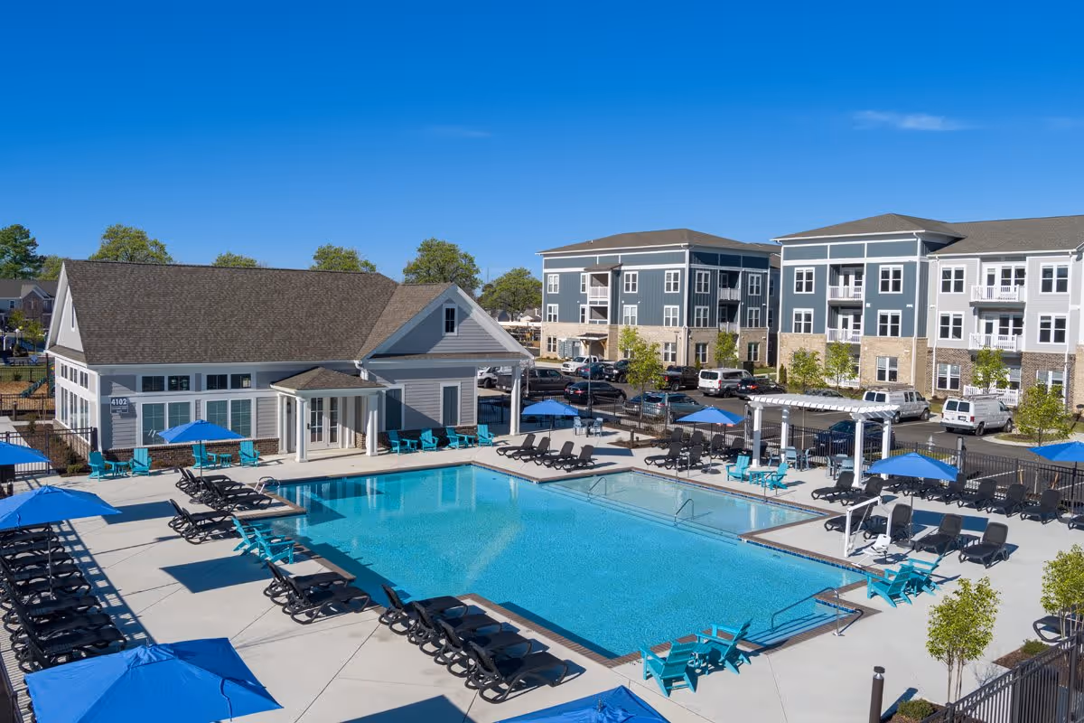 Outdoor swimming pool area at The Compass at Springdale Park with multiple black lounge chairs and blue umbrellas surrounding the pool. There is a small building with a porch and several blue chairs near the pool. In the background, there are multi-story residential buildings and parked cars under a clear blue sky.