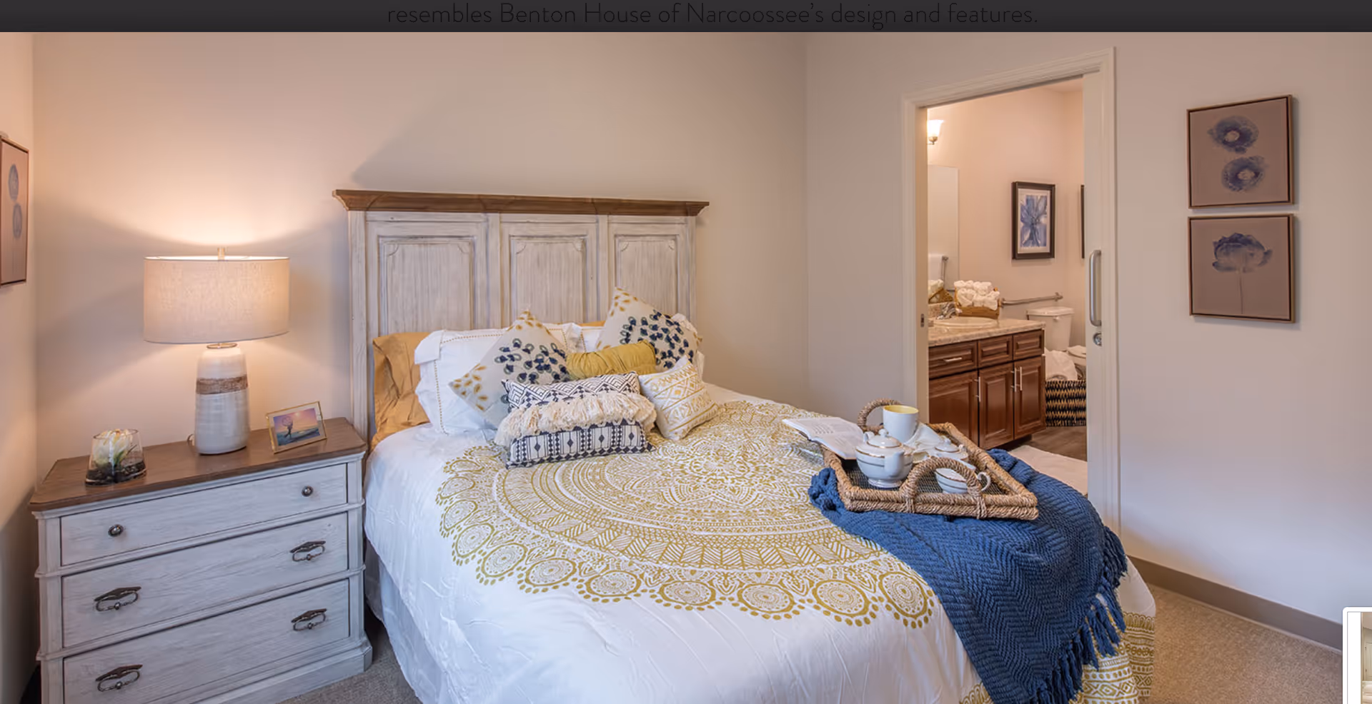 A cozy bedroom with a wooden headboard bed adorned with multiple decorative pillows and a yellow patterned bedspread. A blue throw blanket and a tray with a teapot and cup are placed on the bed. To the left, there is a wooden nightstand with a lamp, a small framed picture, and a decorative item. The room has light-colored walls and carpeted floor. An open doorway reveals a bathroom with a vanity, sink, and toilet.