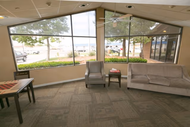 Sunlit lounge with large angled windows showing trees and parked cars, containing a sofa, an armchair, a small table with magazines, and a game table.