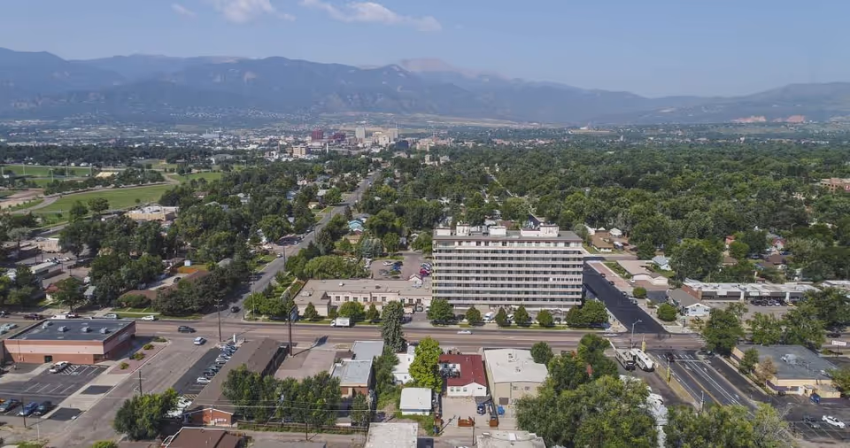 Aerial view of a cityscape with a mix of residential and commercial buildings, tree-lined streets, and a mountain range in the background under a clear blue sky.
