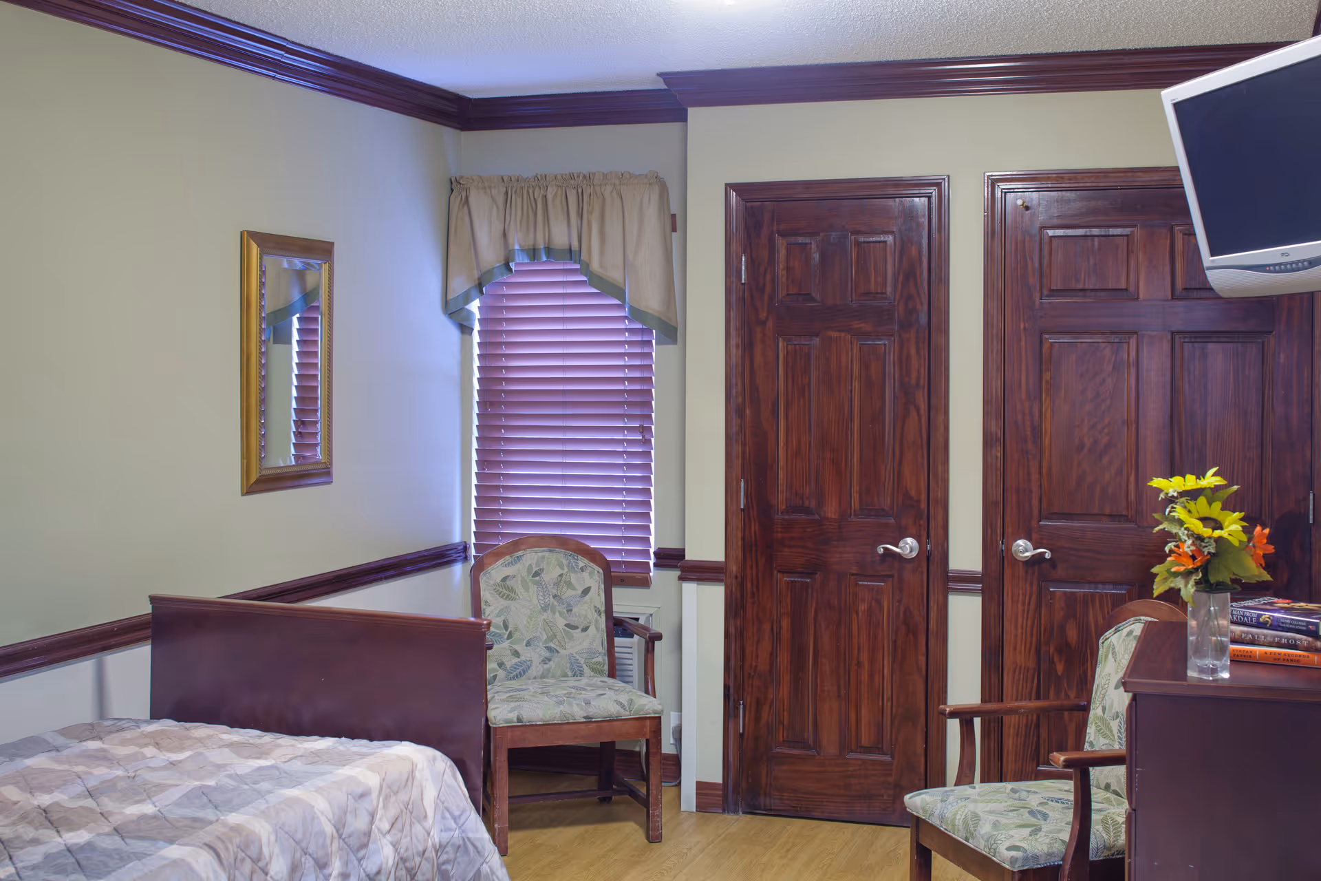 Interior of a bedroom with a bed covered in a patterned quilt, two wooden doors, a window with closed blinds and a valance, two upholstered chairs with floral patterns, a wall-mounted TV, a small table with a vase of flowers and books, and a mirror on the wall.
