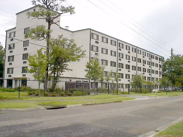 Exterior view of a multi-story residential building with several windows, surrounded by trees and a fenced grassy area, adjacent to a paved road.