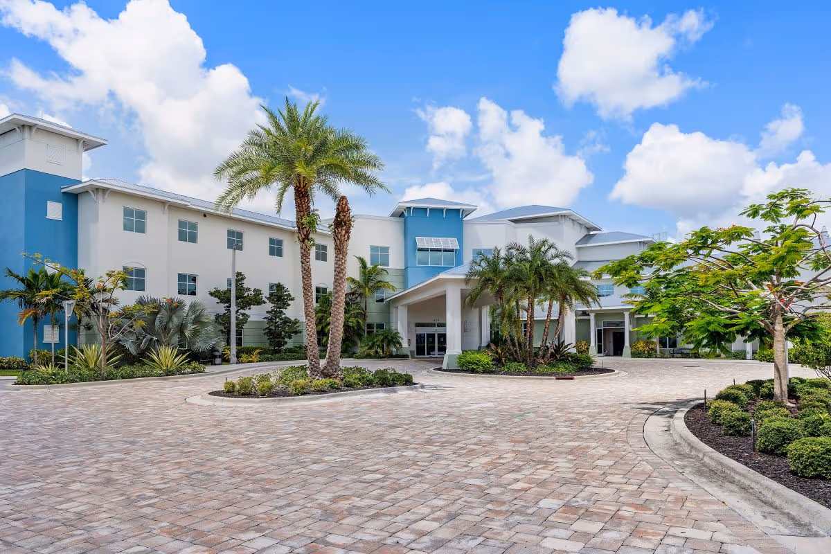 Exterior view of Luxe Senior Living at Jupiter, showing a large, modern building with white and blue walls, surrounded by palm trees and landscaped greenery under a partly cloudy blue sky.
