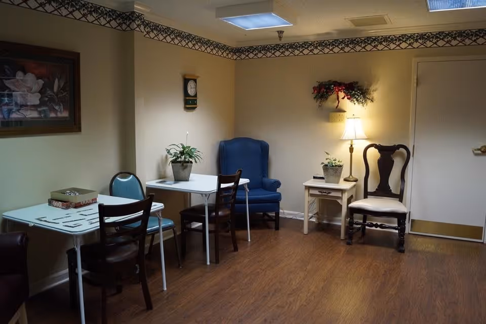 A cozy interior room with two small white tables, each with chairs around them. One table has a puzzle and a box on it. There is a blue upholstered armchair in the corner, a wooden chair with a white cushion next to a small side table with a lamp and a potted plant. The walls are light-colored with a decorative border near the ceiling, and there is a framed floral painting on the wall.