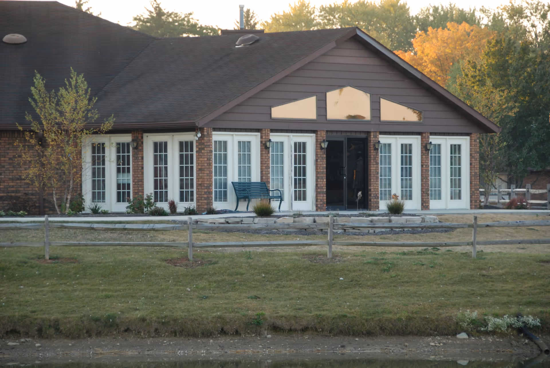 Front view of a single-story brick and wood building with multiple tall French doors, triangular transom windows, a bench on the patio, and a small fence with lawn in front.