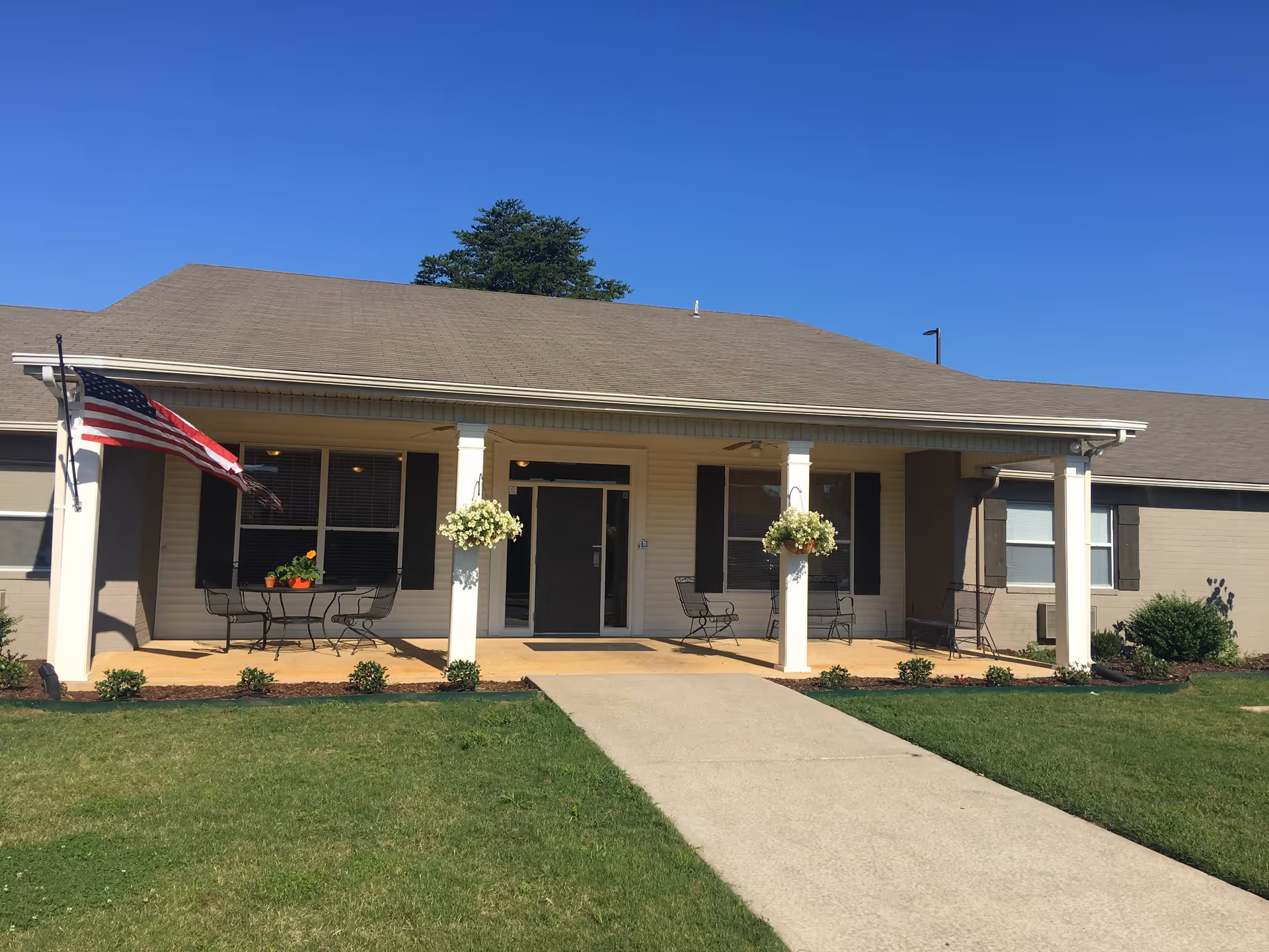 Front exterior view of Limestone Lodge building with a covered porch, white columns, hanging flower baskets, an American flag, outdoor chairs and tables, and a concrete walkway leading to the entrance under a clear blue sky.