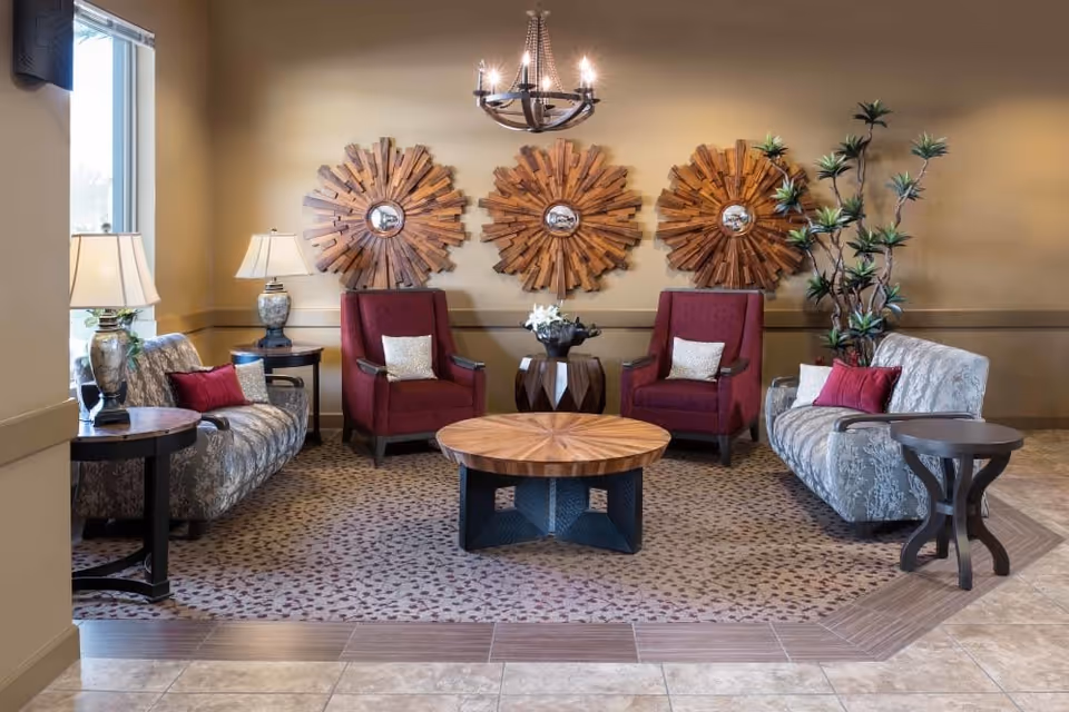 Seating lounge with two patterned sofas, two red armchairs, a round wooden coffee table, lamps, chandelier, and decorative wooden sunburst wall art.
