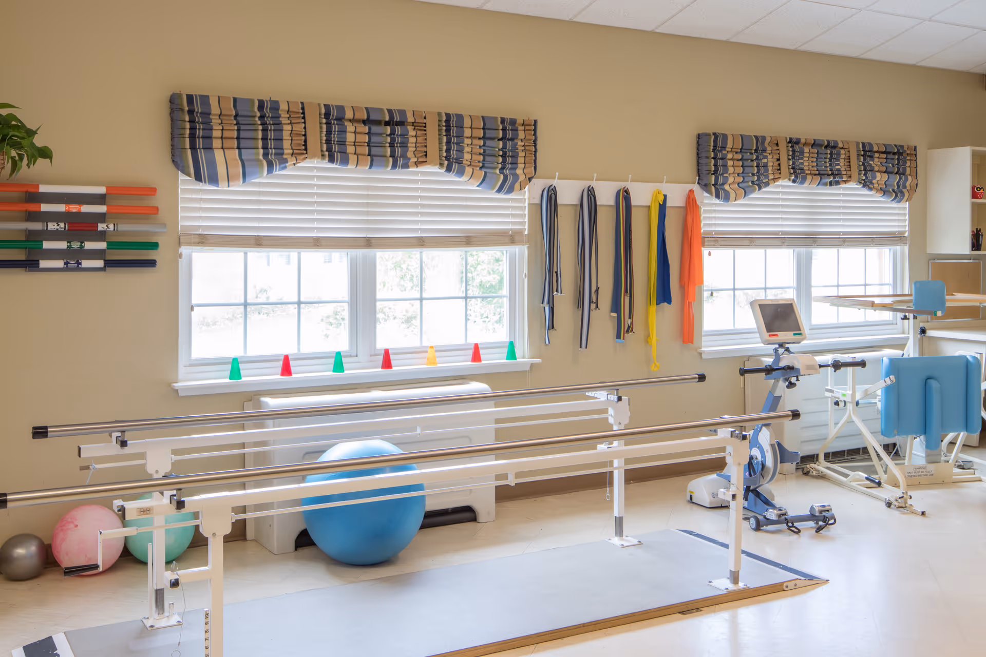 Bright rehabilitation therapy room with parallel walking bars, exercise balls, resistance bands and windows.