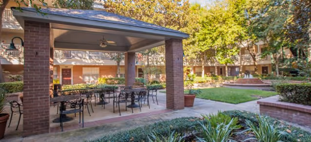 Outdoor courtyard area at Brookdale West University featuring a covered patio with metal tables and chairs, surrounded by greenery, trees, and a multi-story building in the background.