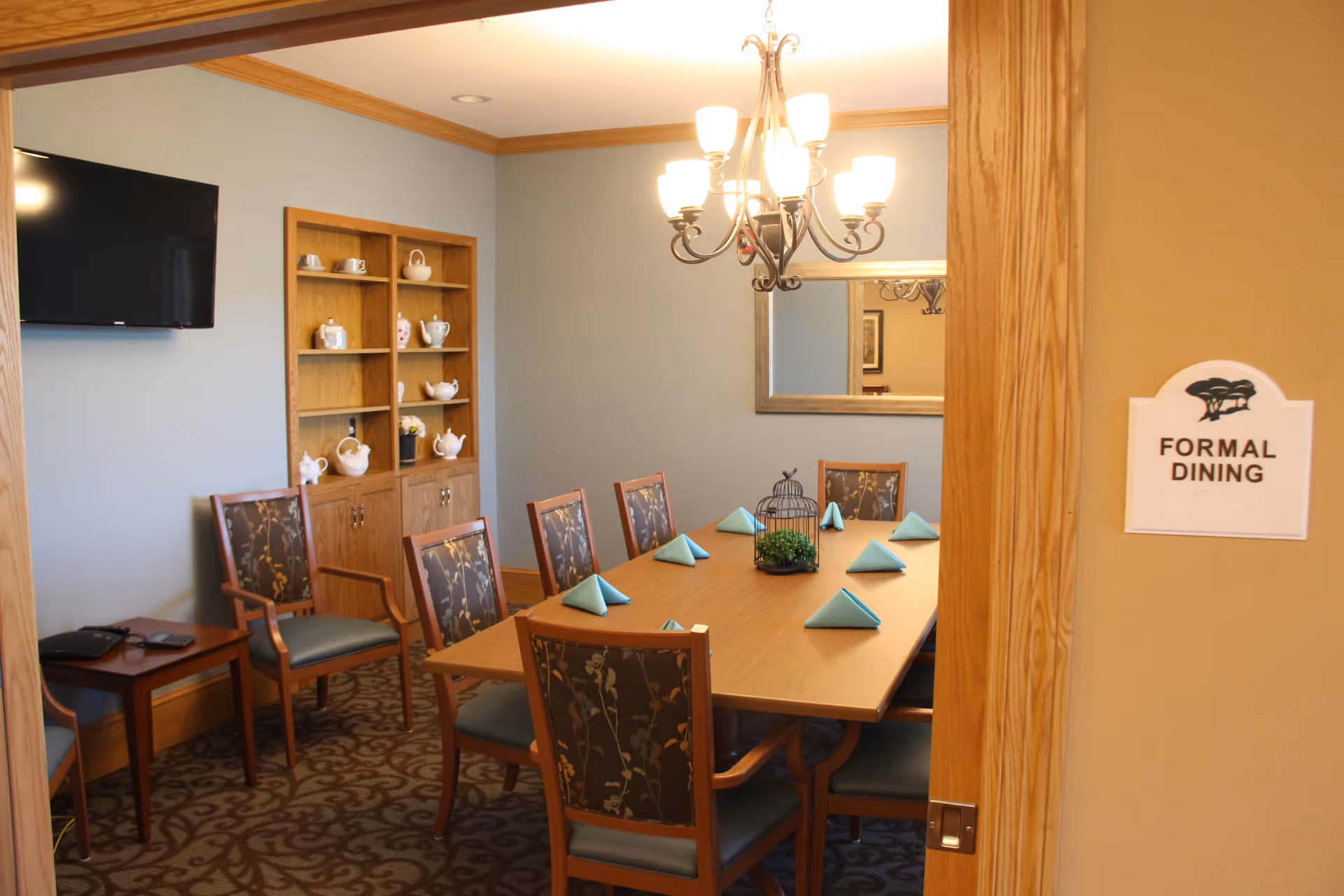 A formal dining room with a rectangular wooden table set with folded blue napkins and a decorative centerpiece. The room has eight upholstered chairs with floral patterns, a chandelier overhead, a wall-mounted flat screen TV, and a wooden cabinet displaying teapots and cups. A sign on the door reads 'FORMAL DINING'.