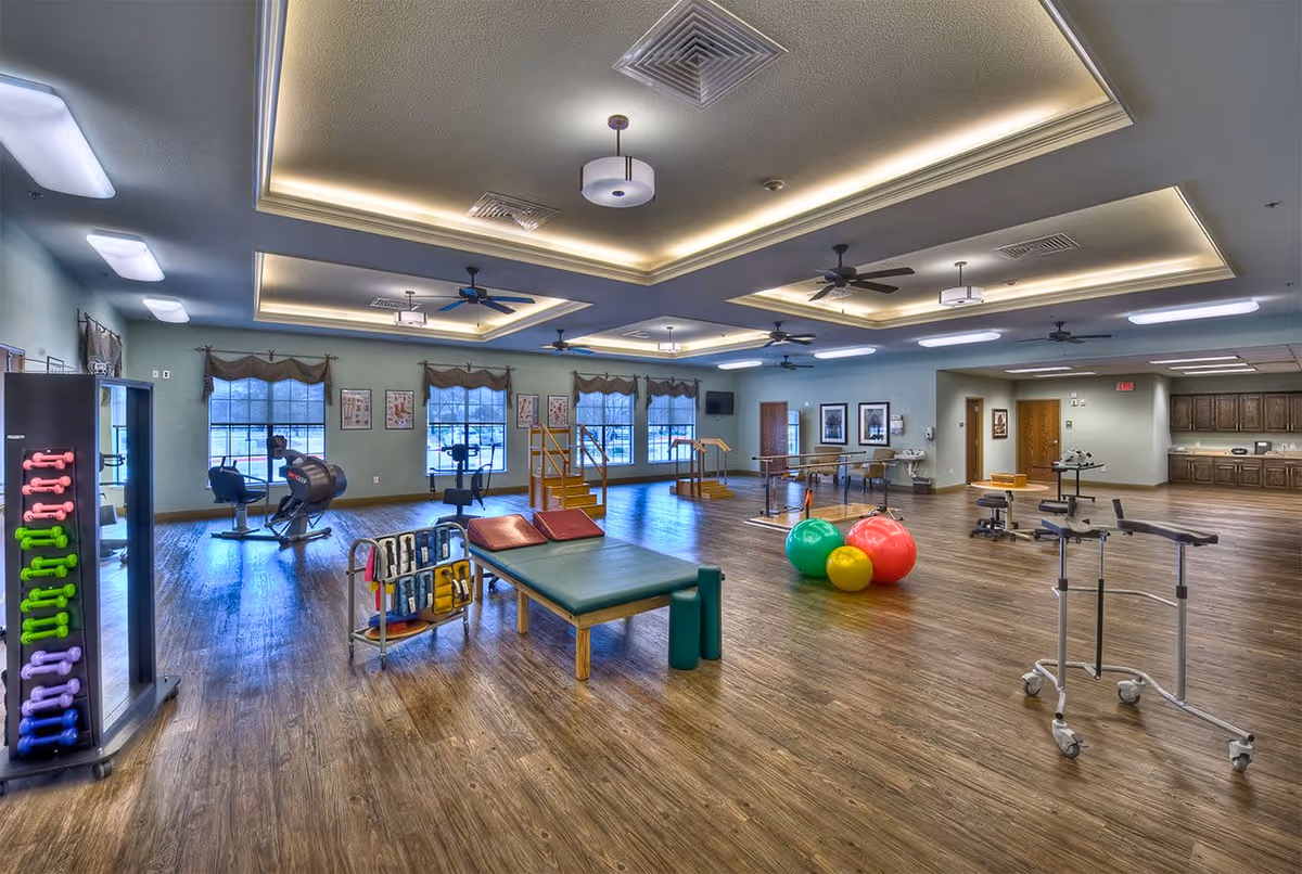 A spacious physical therapy or rehabilitation room with wooden flooring, exercise equipment including colorful dumbbells, therapy tables, exercise balls, parallel bars, and stationary bikes. The room has large windows with valances, recessed ceiling lights, ceiling fans, and a kitchenette area in the back.