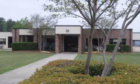 Single-story brick facility entrance with a concrete walkway, lawn, and trees.