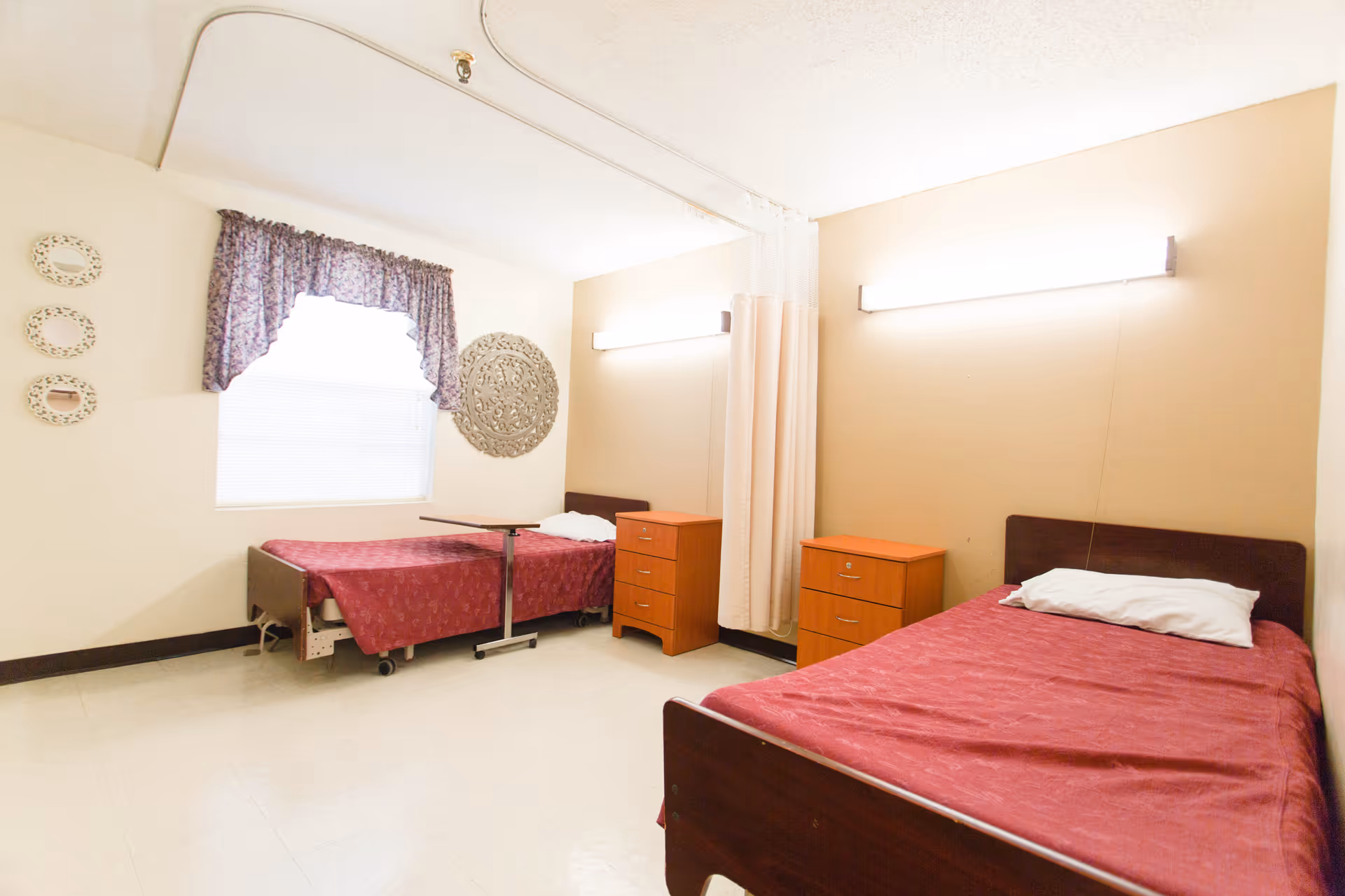 A bright room with two single beds covered in red bedspreads, each with a white pillow. Between the beds are two wooden nightstands with drawers. A curtain divider hangs from the ceiling between the beds. The room has a window with a floral valance and decorative wall hangings. The floor is light-colored and the walls are painted beige and white.