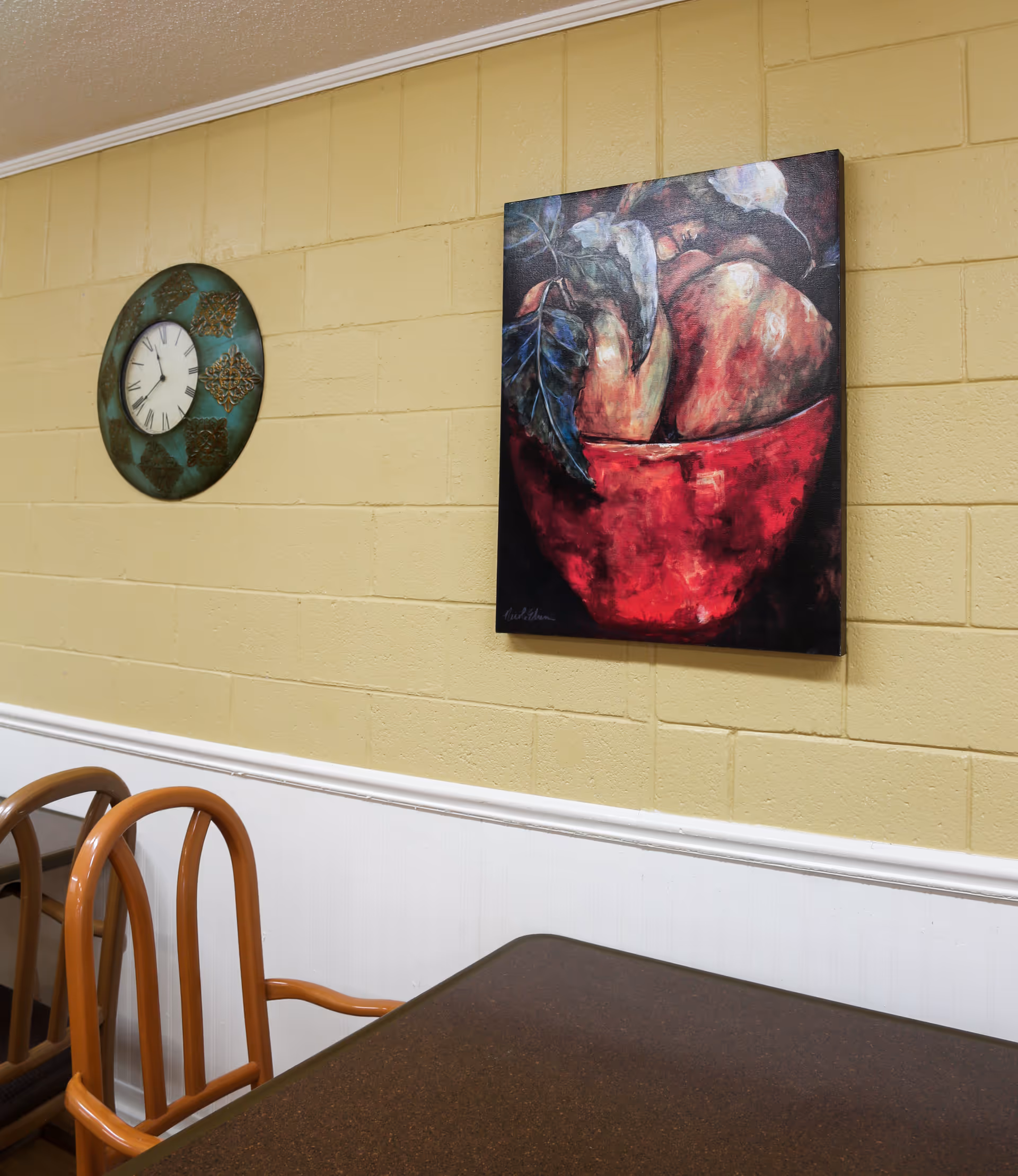 Interior view of a room with a brown table and wooden chairs. On the yellow brick wall, there is a round decorative clock and a painting of a red bowl with fruit and leaves.