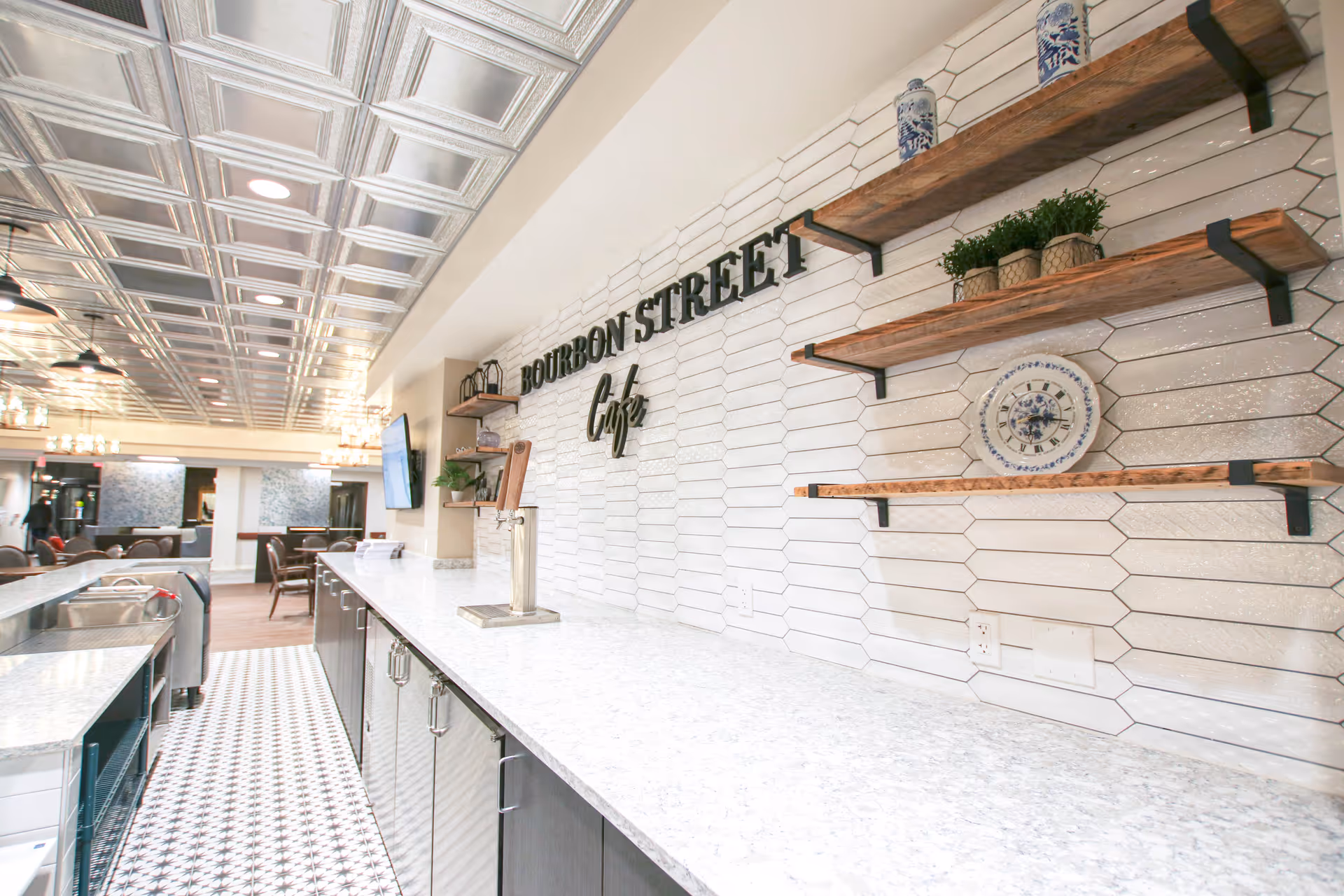 Interior view of the Bourbon Street Café at The Village of Heather Hills featuring a long white marble countertop with dark cabinets underneath, white hexagonal tiled wall with wooden shelves holding decorative items, a silver beverage tap, patterned floor tiles, and a ceiling with metallic square panels and recessed lighting. In the background, there are tables and chairs in a dining area.