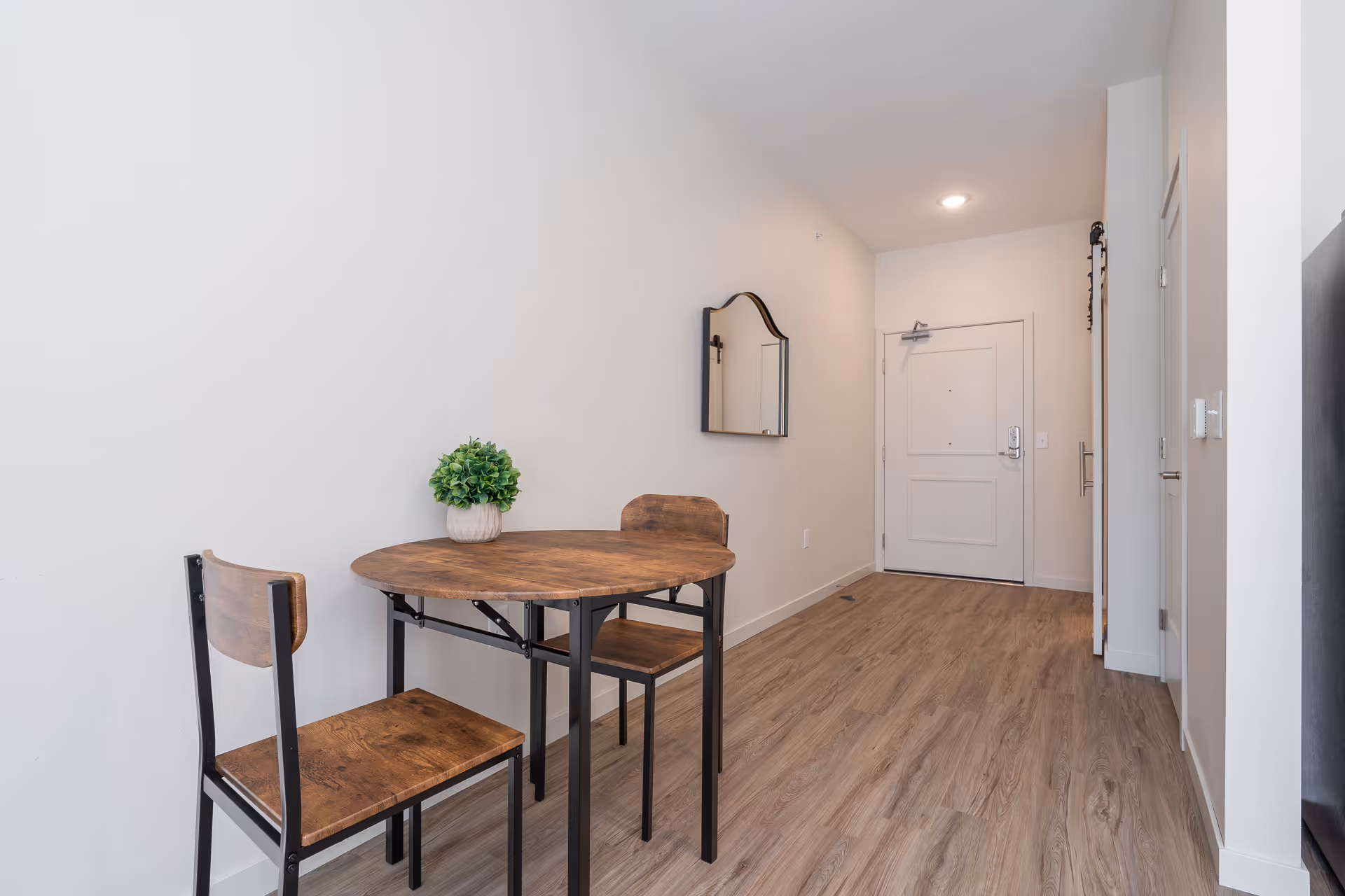 A small dining area with a round wooden table and two matching wooden chairs with black metal legs. A small green potted plant is placed on the table. The area has light-colored walls and wood flooring. A mirror is mounted on the wall near the entrance door in the background.