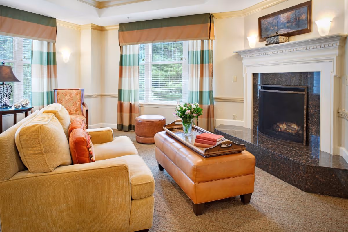 A cozy living room with a beige sofa adorned with orange pillows, a leather ottoman coffee table with a tray holding books and a vase of flowers, a fireplace with a dark marble surround, and large windows with striped curtains letting in natural light.