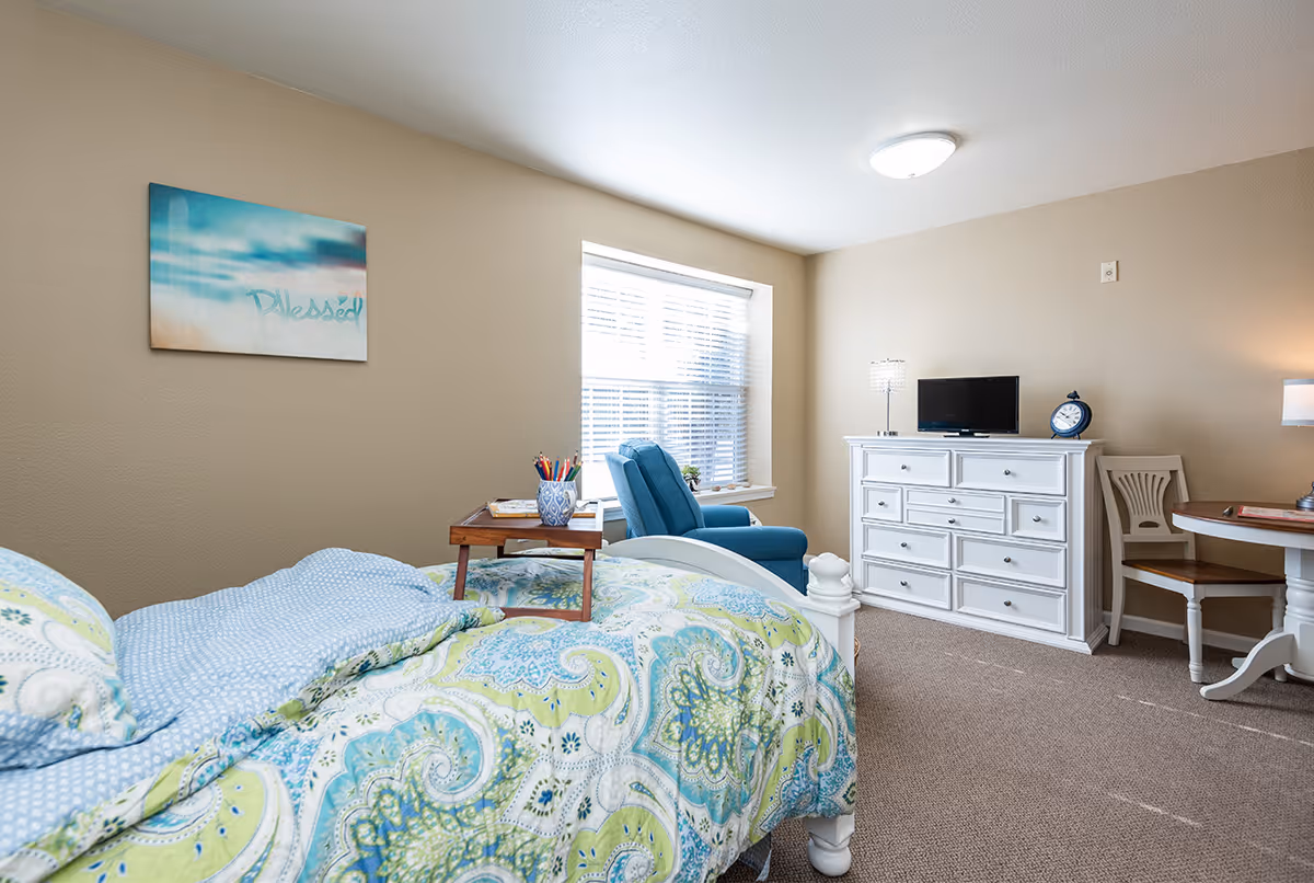 A bright and cozy bedroom in a senior living facility featuring a bed with a colorful paisley-patterned comforter, a small wooden tray with colored pencils on the bed, a blue armchair near a window with blinds, a white dresser with a small TV and a clock on top, and a round table with a chair. A wall art piece with the word 'Blessed' is hung on the beige wall.