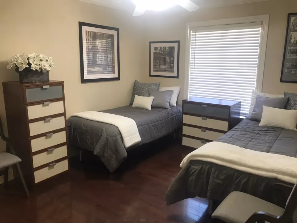 A bedroom with two single beds, each with gray bedding and white throws. Between the beds is a small dresser with three drawers. Another taller dresser with six drawers and a flower arrangement on top is against the left wall. The room has wooden flooring, beige walls, framed pictures on the walls, and a window with closed blinds.