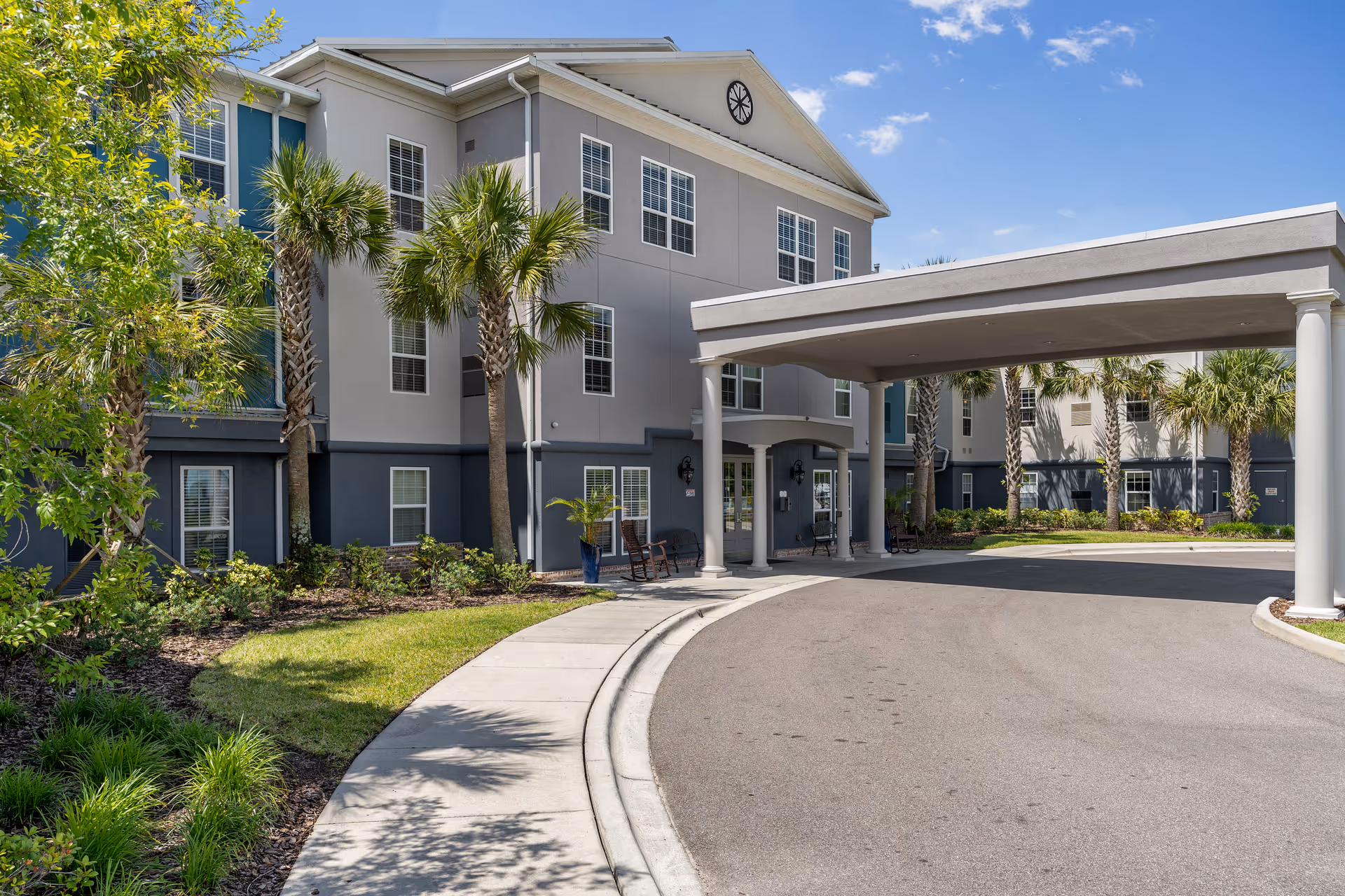 Exterior view of a senior living facility named Gentry Park Orlando, showing a three-story building with multiple windows, palm trees, landscaped greenery, and a covered driveway entrance under a clear blue sky.