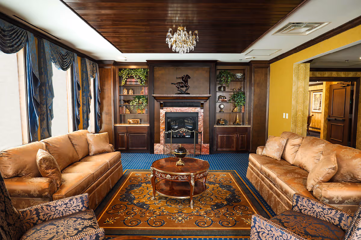 A cozy living room with two beige sofas and two patterned armchairs arranged around a round wooden coffee table on a decorative rug. The room features a fireplace with a dark wood mantel and built-in shelves filled with books and plants. Large windows with blue drapes allow natural light to fill the space, and a chandelier hangs from the wooden ceiling.