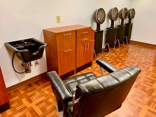 Interior view of a hair salon area with a black salon chair, a black sink for washing hair, a wooden cabinet, and four black hair dryers lined up against a white wall on a parquet floor.