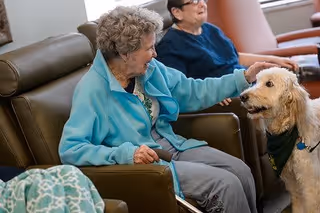 An elderly woman sitting in a brown leather chair petting a light-colored dog wearing a green bandana, with another elderly woman sitting in the background on a chair.