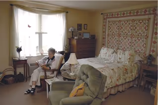 A cozy bedroom with a large bed covered in floral bedding and a quilt hanging on the wall behind it. An elderly woman is sitting in a chair near a window with sheer curtains, reading a book. The room also features a green armchair, a wooden dresser with framed pictures and a lamp, and a small side table with a lamp and other items.