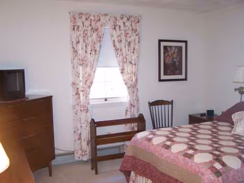 Cozy bedroom with pink floral curtains, quilted bed, dresser with TV, and a window