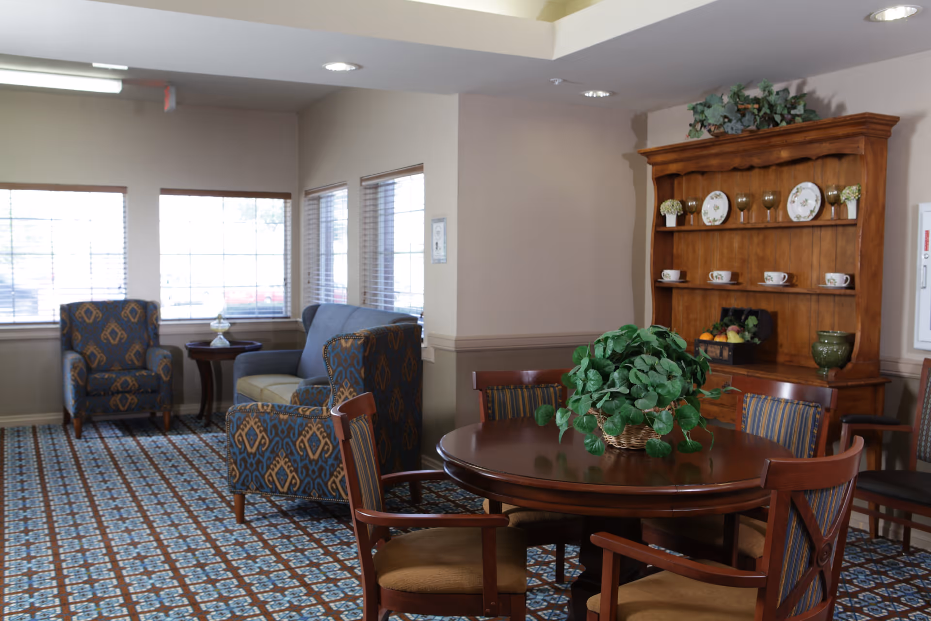 A cozy senior living common area with patterned carpet, a round wooden table with chairs, a green potted plant centerpiece, a wooden hutch displaying decorative plates, cups, and glasses, and a seating area with a blue patterned armchair and a matching loveseat near windows with blinds.