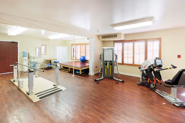 A well-lit rehabilitation or physical therapy room with wooden flooring, featuring parallel bars for walking exercises, a multi-functional exercise machine, a recumbent exercise bike, a large padded platform with a blue exercise ball, and a water cooler near the door. The room has large windows with wooden shutters and a mirror on one wall.
