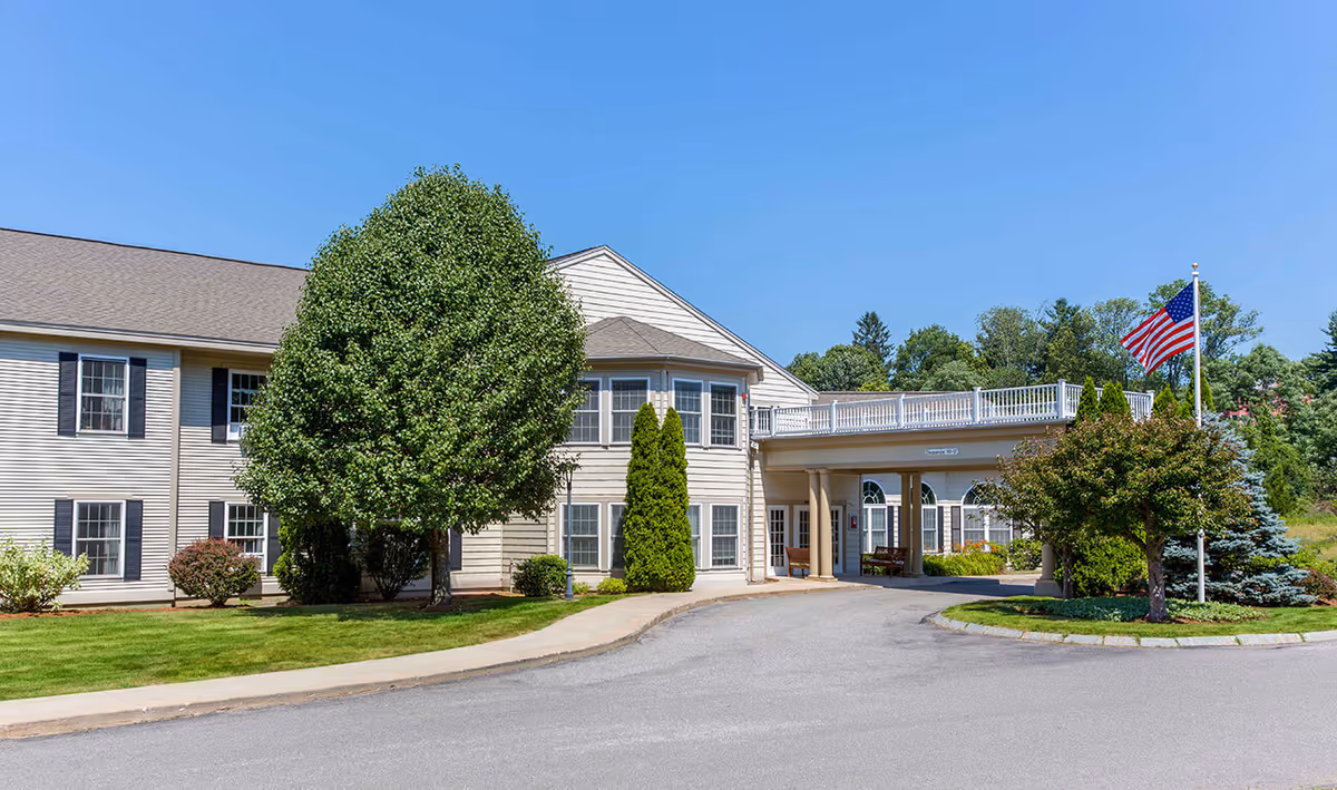 Exterior view of Benchmark at Haverhill Crossings senior living facility showing a two-story building with beige siding, multiple windows, a covered entrance with columns, a large tree, manicured bushes, a lawn, and an American flag on a flagpole under a clear blue sky.